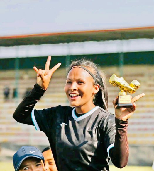 A young woman wearing a soccer uniform holds up a trophy and makes a peace sign with her fingers.