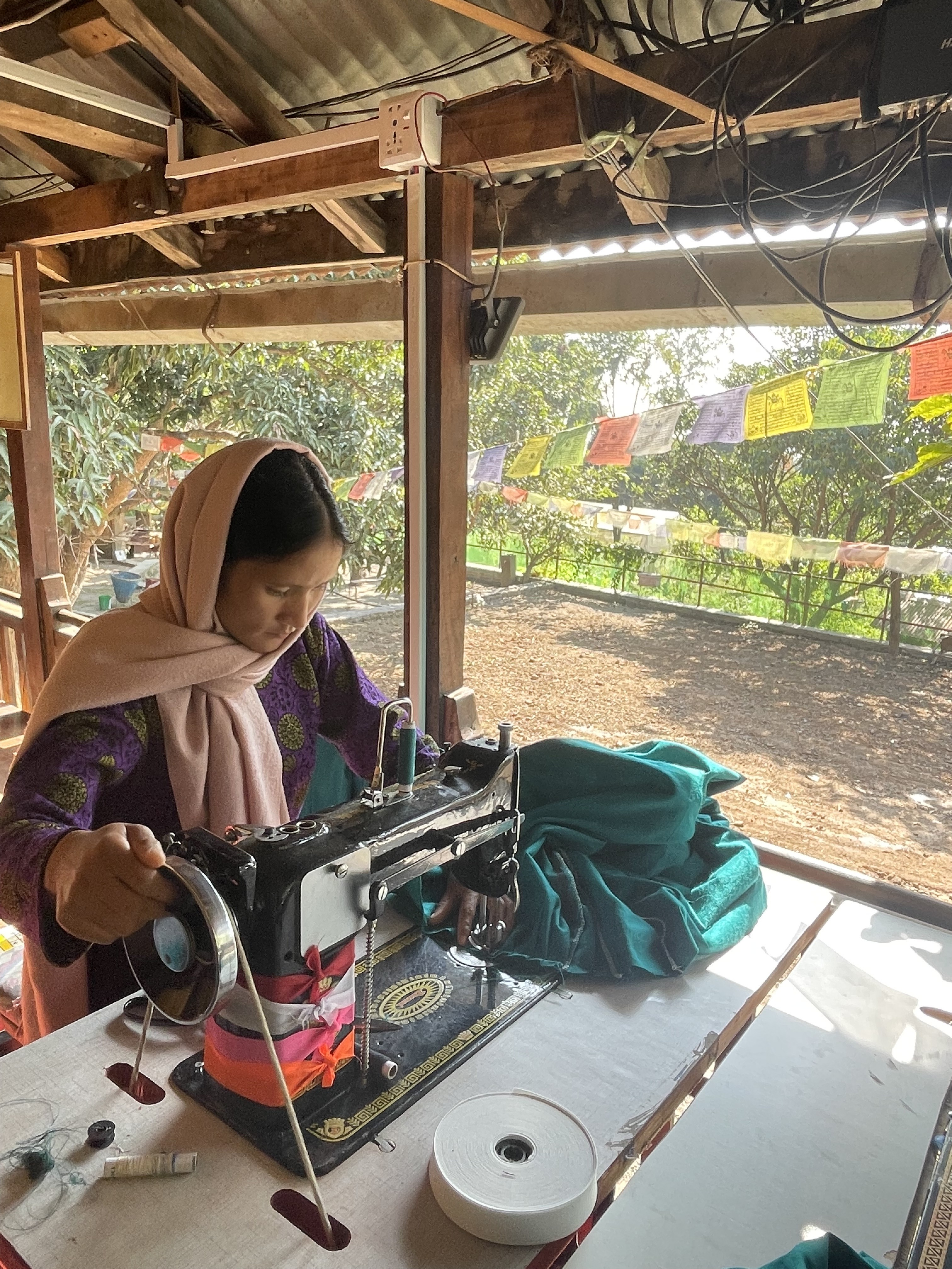 A woman works with a sewing machine on an outdoor porch with a lush tree background.