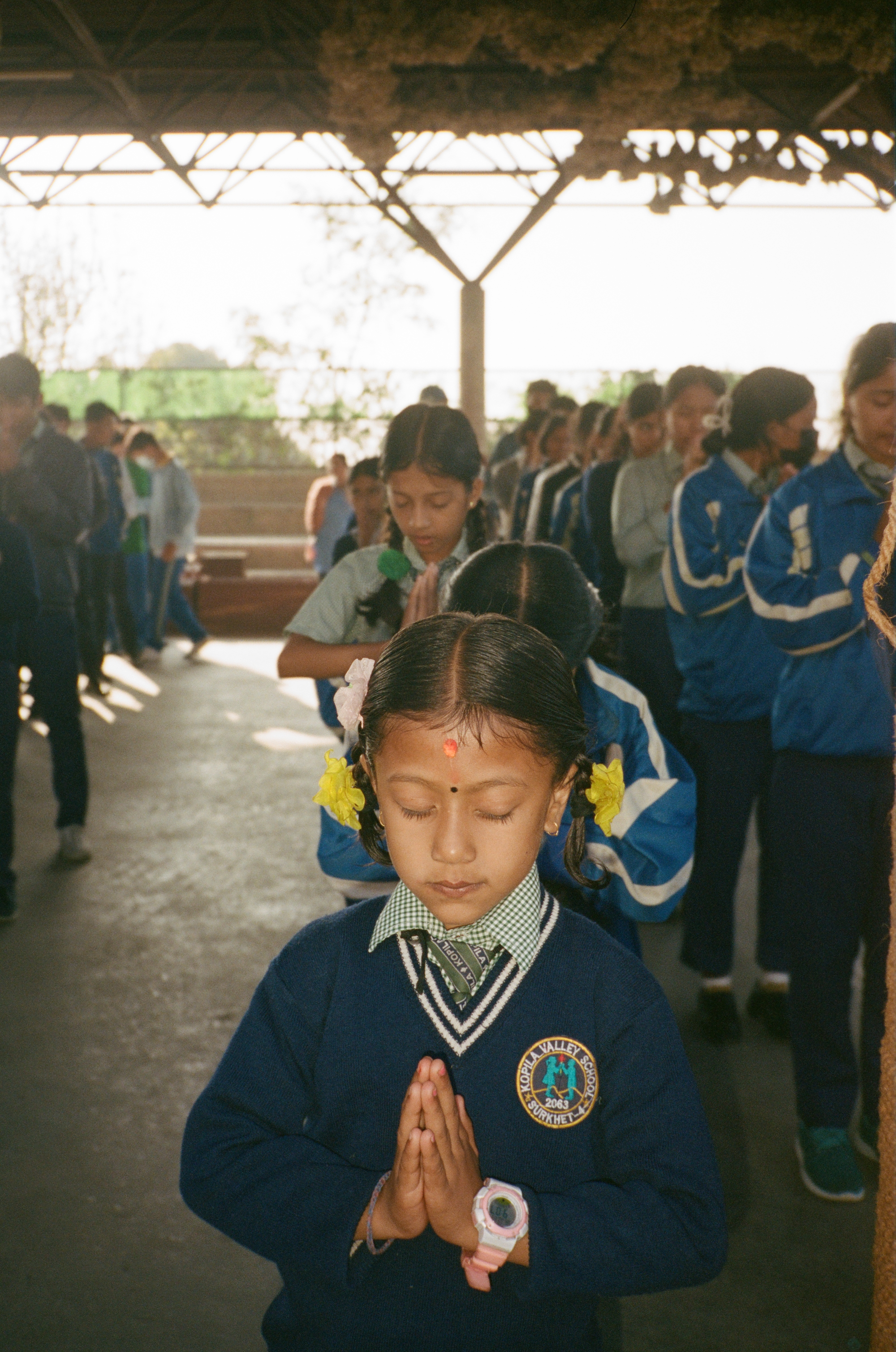A girl in a school uniform closes her eyes in meditation, while standing with a line of children behind her.