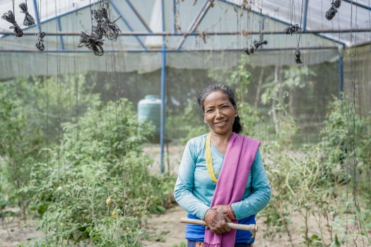 A woman smiles for the camera, standing in front of a greenhouse full of green leafy plants.