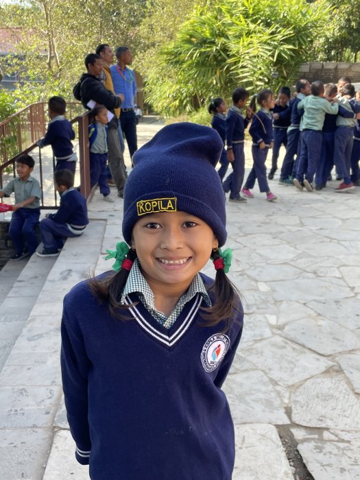 A young girl in a school uniform and stocking cap smiles for the camera.