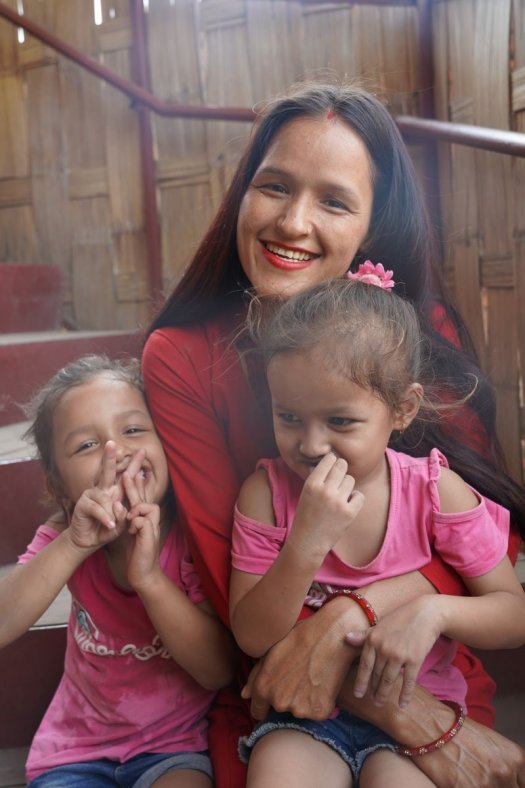 A young woman smiles and holds two young girls on her lap, who are giggling and smiling.